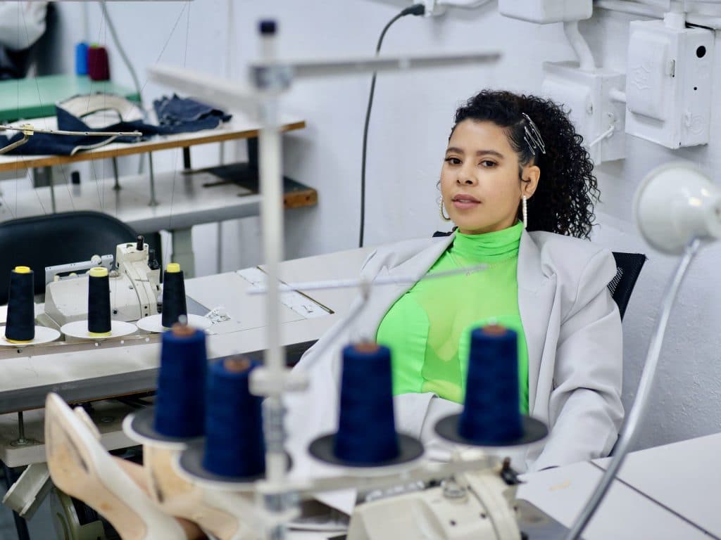 Pale grey oversized blazer over a neon green turtleneck at an industrial sewing bench, cones of navy thread in the foreground.