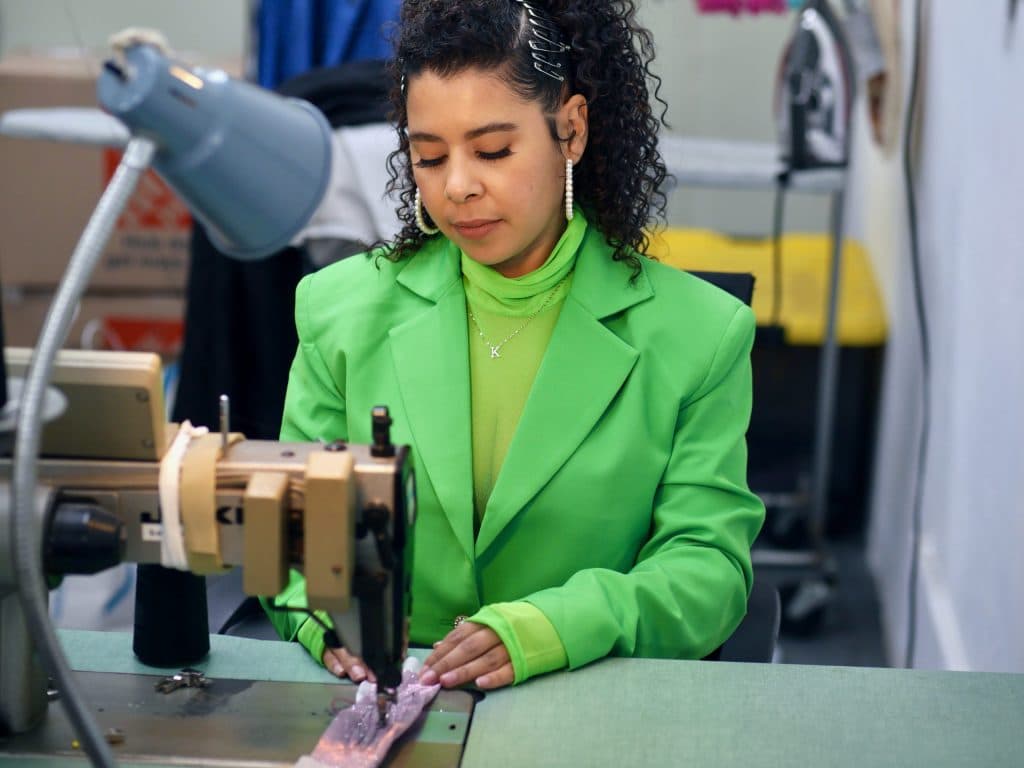 Bright green tailored blazer with padded shoulders, worn while guiding a pale beaded panel under a sewing machine needle.