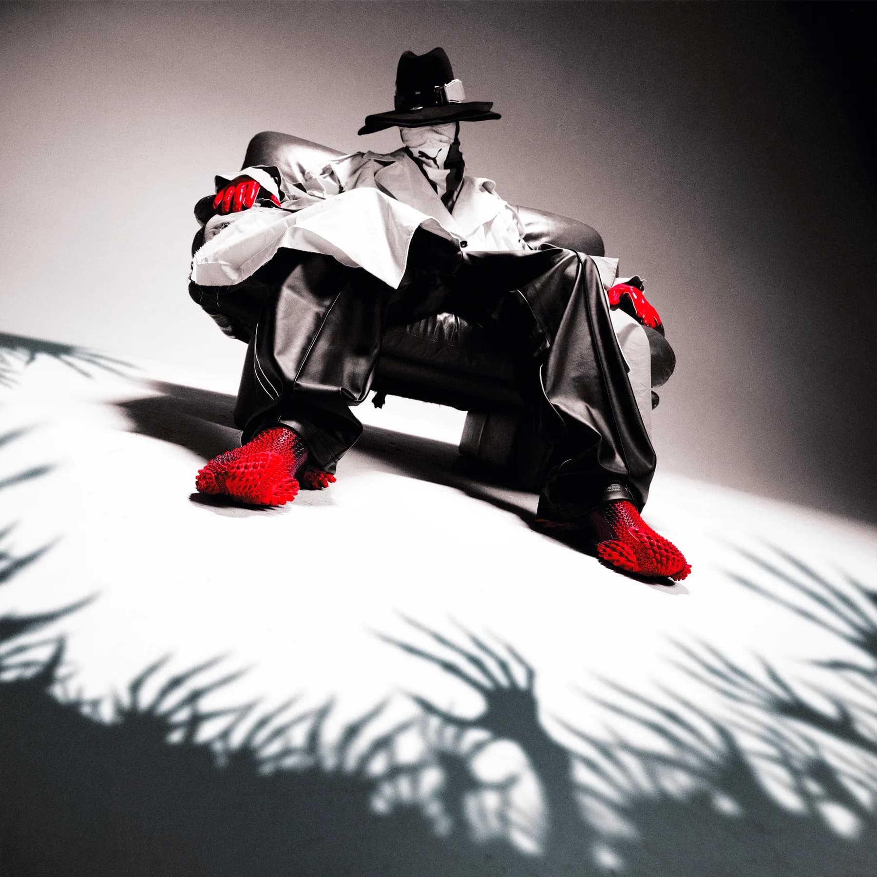 Seated figure in an oversized satin suit and wide brim hat with red spiked shoes, lit so the shadow spreads across the floor like grass.