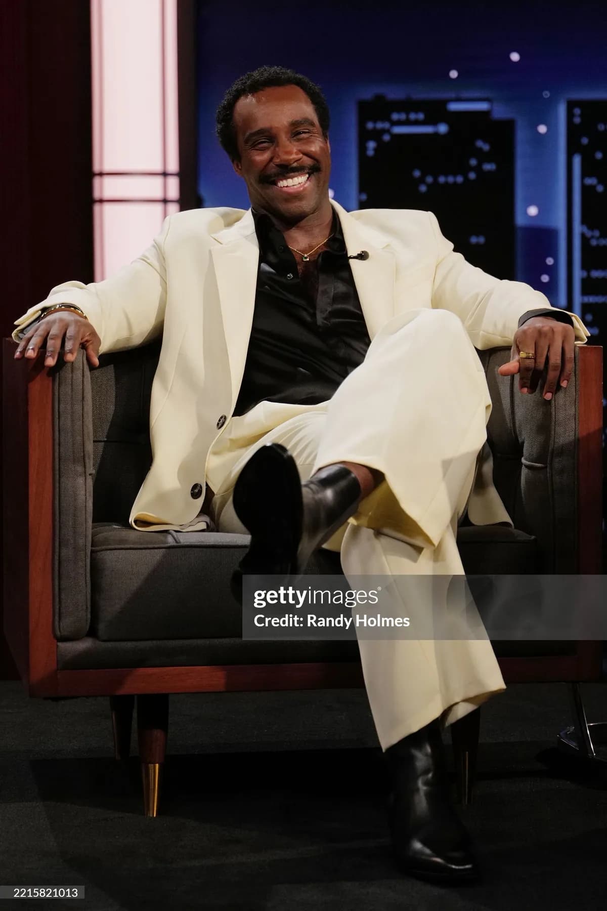 Cream longline coat over a black shirt and black boots, seated in an armchair on a talk show set.