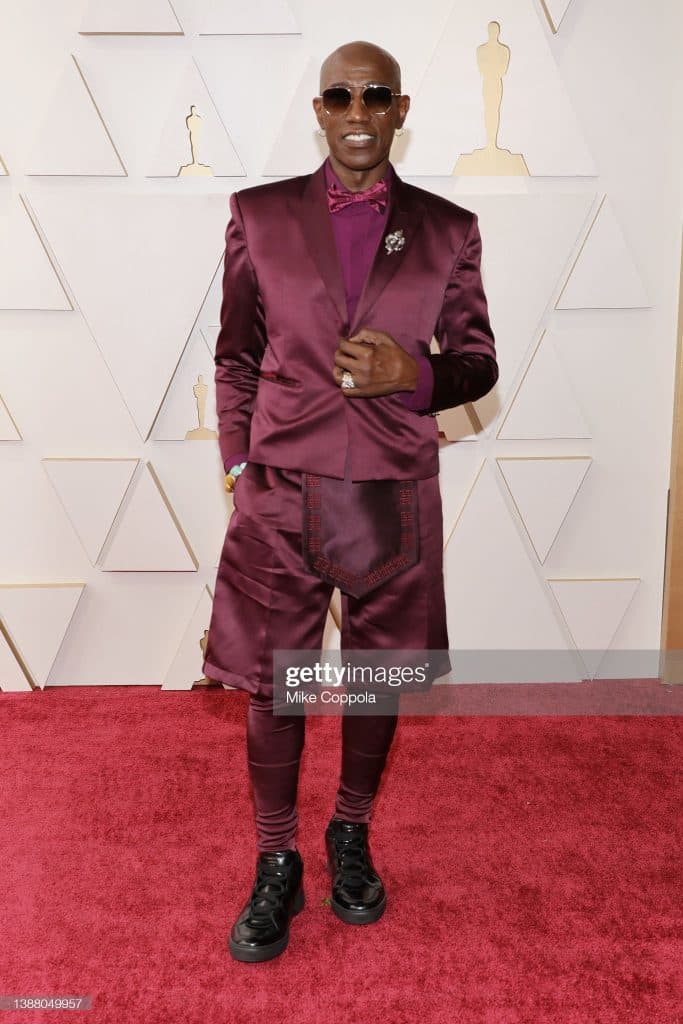 A guest on the awards red carpet in a magenta satin suit and dark sunglasses.
